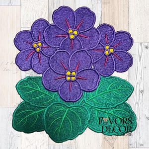 May include: Embroidered violet flower design with purple petals, yellow centres, and red accents. The design includes green leaves and the text "Favors Decor More". The floral design is likely for crafting or home decor.