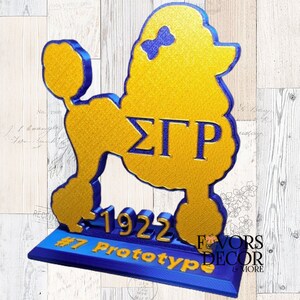 May include: A gold and blue poodle-shaped decorative item. The poodle has a bow on its head and the Greek letters &Sigma;&Gamma;&Rho;. The base reads "1922" and "#7 Prototype". The item is a prototype from Favors Decor & More.