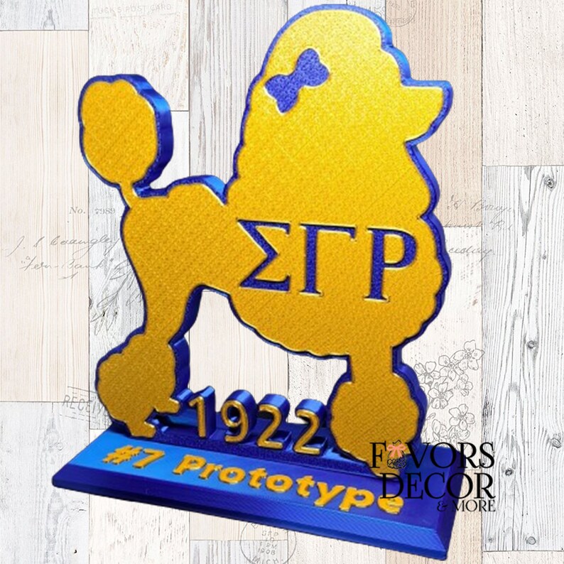 May include: A gold and blue poodle-shaped decorative item. The poodle has a bow on its head and the Greek letters &Sigma;&Gamma;&Rho;. The base reads "1922" and "#7 Prototype". The item is a prototype from Favors Decor & More.