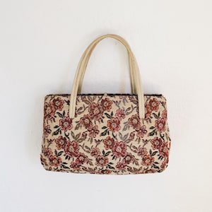 Peut inclure: Sac &agrave; main rectangulaire avec un motif floral dans les tons de rouge, marron et vert sur fond beige. Le sac a deux anses cr&egrave;me. Le sac est fait d'une mati&egrave;re tiss&eacute;e.