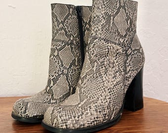 May include: A pair of ankle boots with a snakeskin pattern in shades of gray and beige. The boots have a square toe, a side zipper, and a chunky black heel. The boots are displayed on a wooden table.