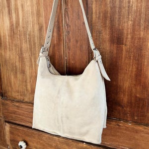 May include: A cream-colored suede shoulder bag with a curved shape and a long, adjustable strap. The bag features a zipper closure and silver-toned hardware. The bag is hanging on a wooden door.