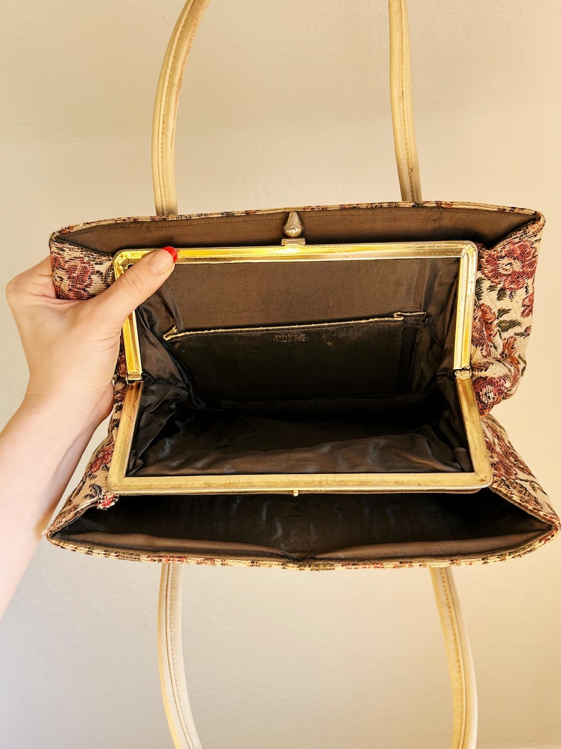 Peut inclure: Sac &agrave; main vintage en tapisserie avec un motif floral rouge, beige et marron. Il est dot&eacute; d'un cadre dor&eacute; et de poign&eacute;es cr&egrave;me. L'int&eacute;rieur est doubl&eacute; de tissu marron fonc&eacute; et le cadre est dot&eacute; d'une fermeture &agrave; fermoir. Le design du sac &eacute;voque une &eacute;poque r&eacute;volue.