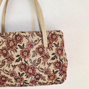 Peut inclure: Sac &agrave; main vintage avec un motif floral en tapisserie. Le sac pr&eacute;sente un fond cr&egrave;me avec des fleurs bordeaux et rouille et des feuilles vertes. Il a des poign&eacute;es cr&egrave;me.