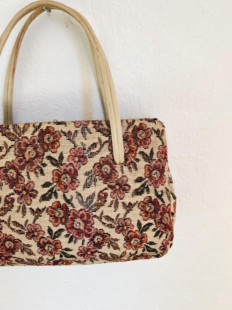Peut inclure: Sac &agrave; main vintage avec un motif floral en tapisserie. Le sac pr&eacute;sente un fond cr&egrave;me avec des fleurs bordeaux et rouille et des feuilles vertes. Il a des poign&eacute;es cr&egrave;me.