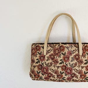 Peut inclure: Un sac &agrave; main rectangulaire avec un motif floral en tapisserie. Le sac pr&eacute;sente un fond beige avec des fleurs bordeaux et roses et des feuilles vertes. Il a deux poign&eacute;es beiges.