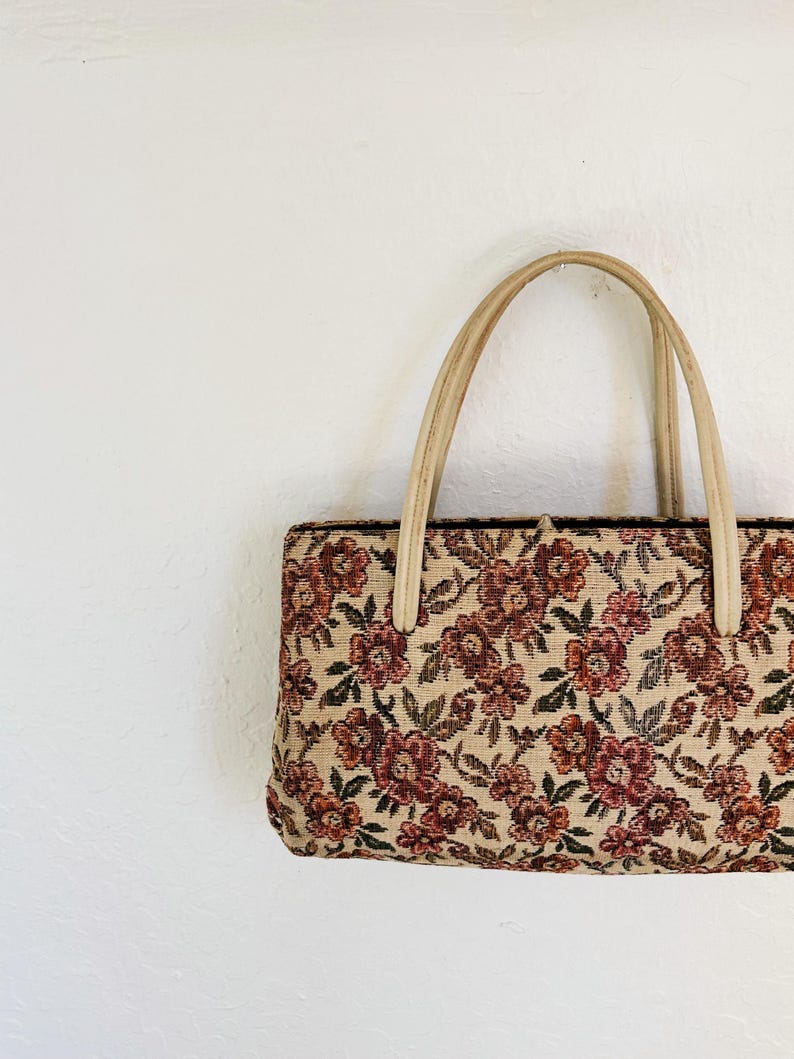 Peut inclure: Un sac &agrave; main rectangulaire avec un motif floral en tapisserie. Le sac pr&eacute;sente un fond beige avec des fleurs bordeaux et roses et des feuilles vertes. Il a deux poign&eacute;es beiges.