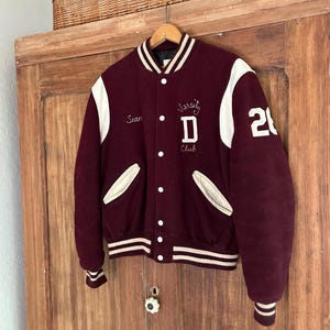 May include: A maroon varsity jacket with white leather sleeves, featuring a white striped collar, cuffs, and hem. The front displays the embroidered text "Sean" and "Varsity Club" with a letter "D", while the number "21" is on the sleeve.