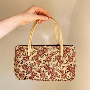 Peut inclure: Un sac &agrave; main rectangulaire en tapisserie avec un motif floral dans les tons de rouge, marron et beige. Le sac a deux poign&eacute;es beiges. Le sac est tenu par une main.