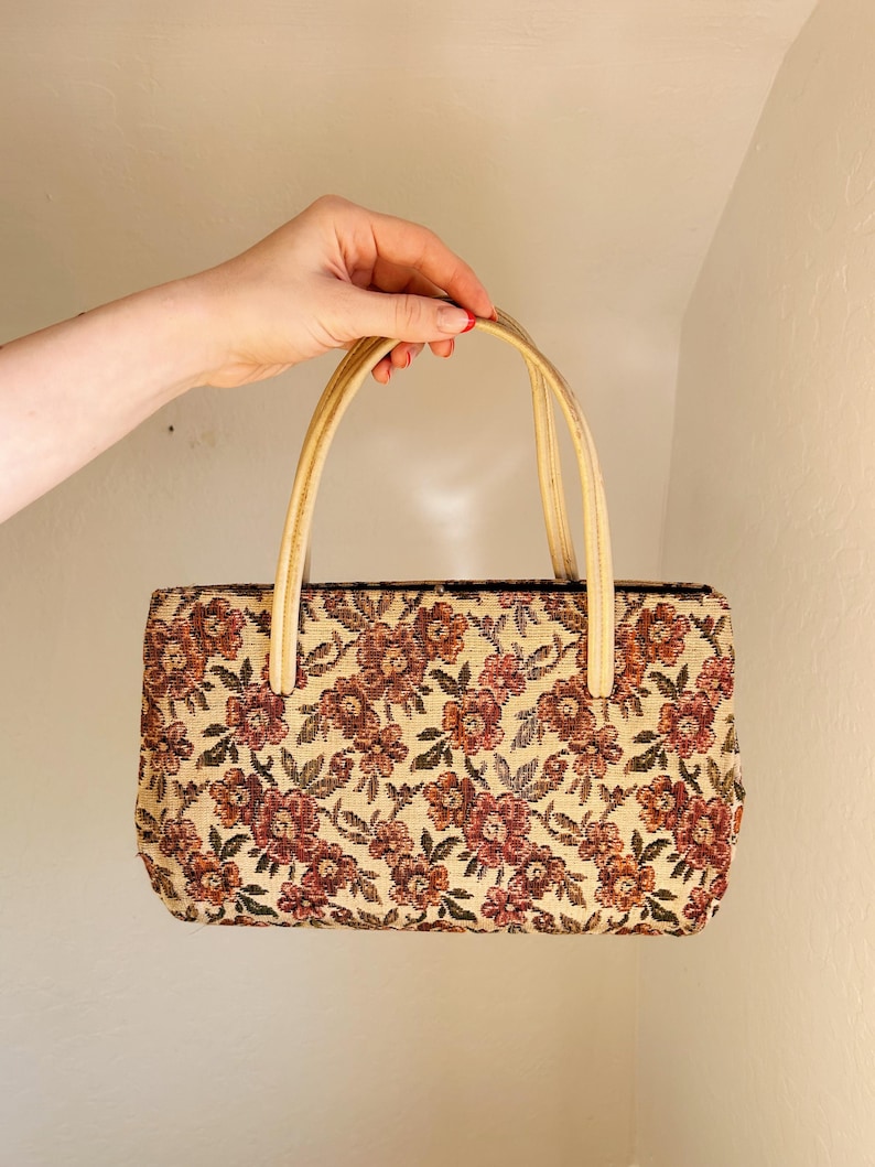 Peut inclure: Un sac &agrave; main rectangulaire en tapisserie avec un motif floral dans les tons de rouge, marron et beige. Le sac a deux poign&eacute;es beiges. Le sac est tenu par une main.