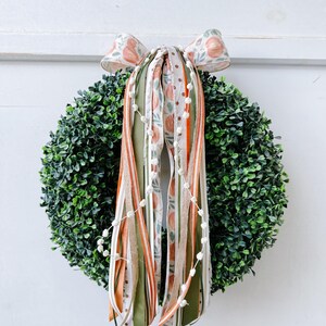 May include: A round, green boxwood wreath adorned with a bow and cascading ribbons. The bow features a peach and floral pattern, while the ribbons include stripes and a dotted design. A string of white beads hangs from the center.