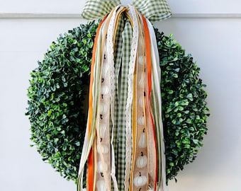 May include: A round, green wreath with a gingham bow and colorful ribbons. The ribbons are adorned with white pumpkins and lace. The wreath is hanging on a white surface.