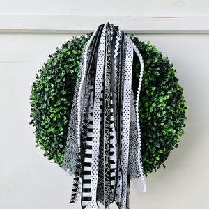 May include: A round, green wreath with a variety of black and white ribbons hanging from it. The ribbons have different patterns, including polka dots, stripes, and spiderwebs. The wreath is hanging on a white background.