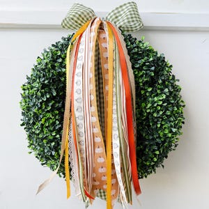 May include: A round, green wreath with a bow made of green and white checkered fabric. The wreath is adorned with various ribbons in orange, white, yellow, and brown, some with pumpkin patterns. The wreath is hanging on a white surface.