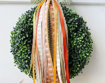May include: A round, green wreath with a bow made of green and white checkered fabric. The wreath is adorned with various ribbons in orange, white, yellow, and brown, some with pumpkin patterns. The wreath is hanging on a white surface.