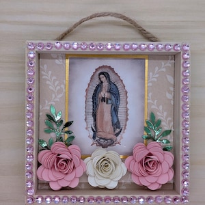 May include: A framed religious artwork featuring an image of the Virgin Mary. The wooden frame is adorned with pink rhinestones and paper flowers. A jute rope is attached for hanging. The artwork has a beige background.