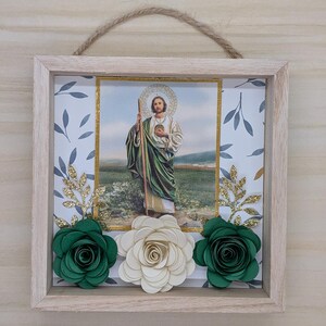 May include: Square framed artwork featuring an image of a religious figure with a halo, holding a staff. The frame is decorated with green and white paper flowers, gold glitter leaves, and a rope hanger.
