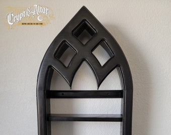 May include: Black wooden shelf unit with a gothic arch design. The shelf has three shelves and is painted black. The shelf is mounted on a white wall.