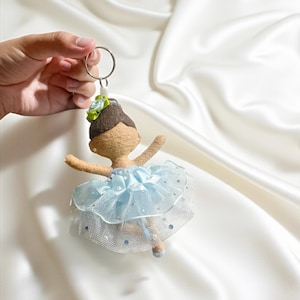 May include: A handmade ballerina keychain. The doll has a tan body, brown hair, and a light blue tutu with silver accents. A small green and blue flower adorns the top of the head. The keychain is attached to a silver ring.