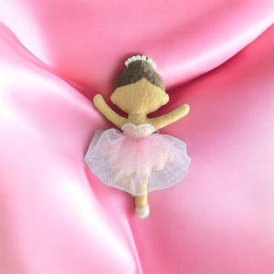 May include: A handcrafted ballerina doll with a yellow face and arms, brown hair, and a pink tutu. The doll features a white beaded tiara and is set against a pink satin backdrop. The tutu is made of delicate white mesh.