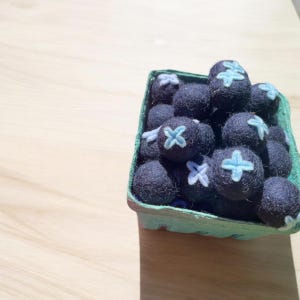 May include: A punnet of dark blue felt blueberries, each with a light blue cross design. The punnet is green and sits on a light brown wooden surface. The blueberries are made of felt.