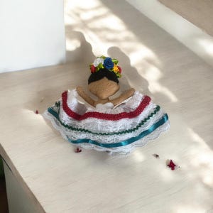 May include: A handmade doll dressed in a traditional Mexican dress. The doll has a brown face, black hair, and a floral crown. The white dress is trimmed with red, green, and blue ribbons and lace.