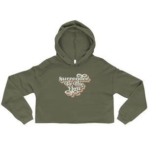 May include: Olive green cropped hoodie with a drawstring hood and long sleeves. The front features the text "Surrender To The Flow" in a retro font with green, orange, and white colors. A casual, comfortable top.
