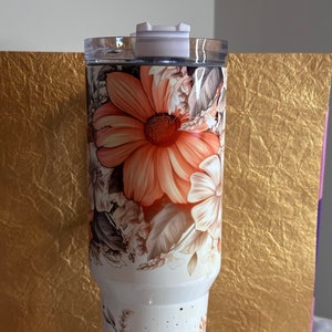 Puede incluir: Un vaso blanco con un dise&ntilde;o floral con flores de color naranja, marr&oacute;n y blanco. El vaso tiene una tapa y est&aacute; hecho de acero inoxidable.