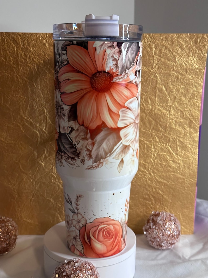 Puede incluir: Un vaso blanco con un dise&ntilde;o floral con flores de color naranja, marr&oacute;n y blanco. El vaso tiene una tapa y est&aacute; hecho de acero inoxidable.