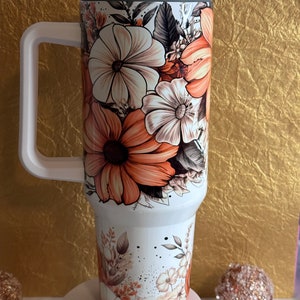 Puede incluir: Un vaso de acero inoxidable blanco con un dise&ntilde;o floral con flores de color naranja, marr&oacute;n y blanco. El vaso tiene una tapa y un asa.