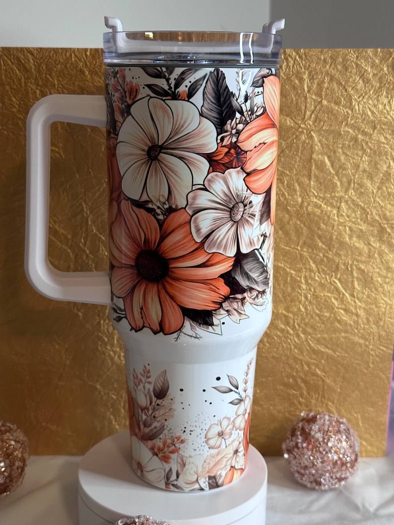 Puede incluir: Un vaso de acero inoxidable blanco con un dise&ntilde;o floral con flores de color naranja, marr&oacute;n y blanco. El vaso tiene una tapa y un asa.
