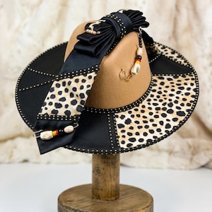 Puede incluir: Sombrero fedora con copa marrón y ala negra. El ala presenta un estampado de leopardo y tachuelas plateadas. Una cinta negra con diseño de leopardo y cuentas decorativas añade un toque único. El sombrero se exhibe en un soporte de madera.