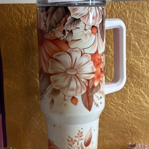 Puede incluir: Taza de viaje blanca con un dise&ntilde;o floral con flores de color naranja, marr&oacute;n y blanco. La taza tiene una tapa transparente y un asa.