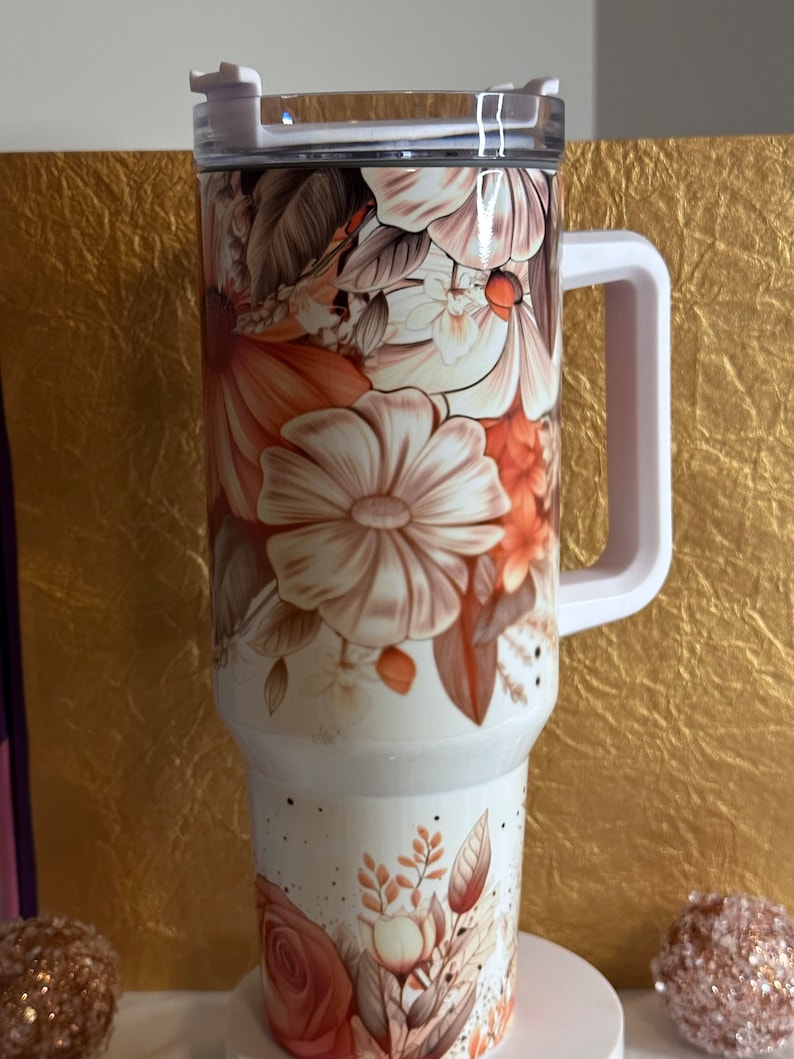 Puede incluir: Taza de viaje blanca con un dise&ntilde;o floral con flores de color naranja, marr&oacute;n y blanco. La taza tiene una tapa transparente y un asa.