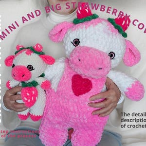 Könnte beinhalten: Zwei gehäkelte Erdbeerkühe, eine große und eine kleine, werden gezeigt. Die größere Kuh ist rosa und weiß mit einem roten Herz und Erdbeerdetails. Die kleinere Kuh hat ähnliche Merkmale. Der Text auf dem Bild lautet "MINI AND BIG STRAWBERRY COWS" und "CROCHET PATTERN."