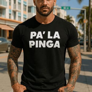 May include: Black crew neck t-shirt with the white text "PA' LA PINGA". The person is standing outdoors with arms at their sides. The person has tattoos on both arms.