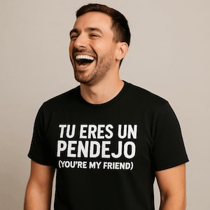 May include: Black crew neck t-shirt with white text that reads "TU ERES UN PENDEJO (YOU'RE MY FRIEND)". A person is wearing the shirt and laughing.