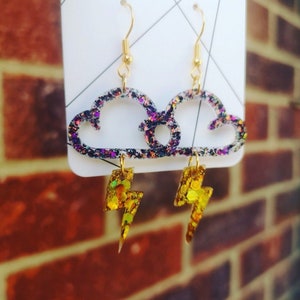 May include: A pair of gold glitter cloud earrings with dangling lightning bolt charms. The clouds are iridescent and have a purple and black glitter finish.