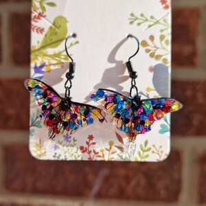 May include: A pair of colourful butterfly earrings with black metal hooks. The butterflies are made of clear resin with a rainbow of colours embedded inside.