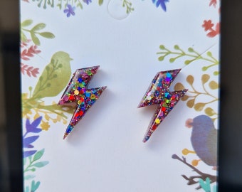 May include: A pair of small, colorful lightning bolt shaped earrings. The earrings are made of resin and have a glittery finish. The glitter is a mix of red, pink, blue, green, and gold.