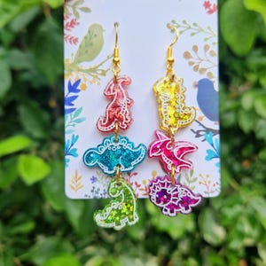 May include: A pair of colorful dinosaur-themed earrings. Each earring features four different dinosaur shapes in red, blue, yellow, pink, and green with gold-colored hooks. The dinosaurs have glittery accents and are attached to each other.
