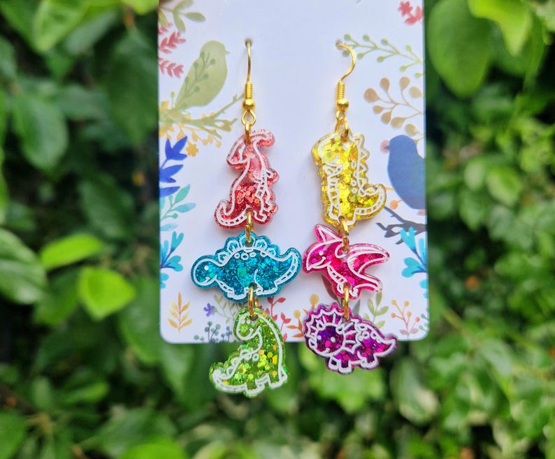 May include: A pair of colorful dinosaur-themed earrings. Each earring features four different dinosaur shapes in red, blue, yellow, pink, and green with gold-colored hooks. The dinosaurs have glittery accents and are attached to each other.