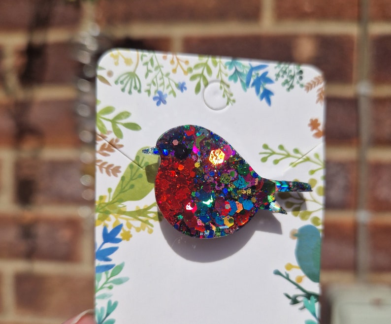 Peut inclure: Une broche en forme de rouge-gorge en paillettes rouges, bleues et vertes. Le rouge-gorge est en r&eacute;sine et a une finition brillante et iris&eacute;e.