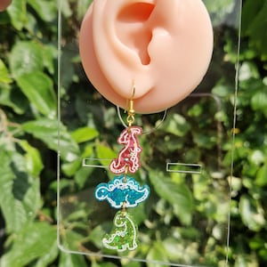 May include: A pair of dangling earrings featuring three dinosaur shapes in red, blue, and green. The earrings have gold-colored hooks and are displayed against a background of green foliage.