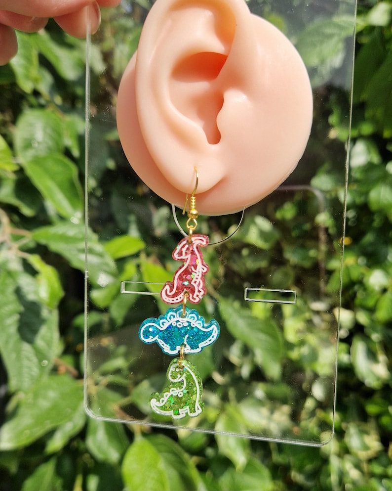 May include: A pair of dangling earrings featuring three dinosaur shapes in red, blue, and green. The earrings have gold-colored hooks and are displayed against a background of green foliage.