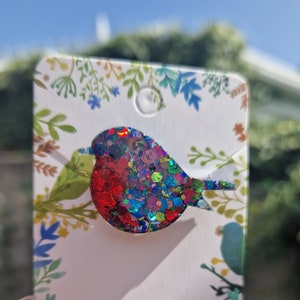 Peut inclure: Une &eacute;pingle de robin en paillettes rouge, bleu et vert. L'&eacute;pingle est sur une carte blanche avec un motif floral.