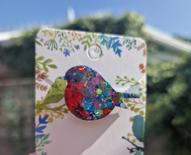 Peut inclure: Une &eacute;pingle de robin en paillettes rouge, bleu et vert. L'&eacute;pingle est sur une carte blanche avec un motif floral.