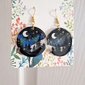 May include: Round dangle earrings featuring a night sky scene with a white crescent moon, gold stars, and a blue and green aurora borealis effect over a black silhouette of pine trees. The earrings have gold-toned metal fishhook ear wires.
