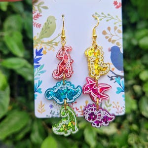 May include: Dangle earrings featuring colorful dinosaur shapes in red, yellow, blue, pink, and green with gold-colored hooks. The earrings have a glittery finish and are displayed on a white card with a floral design.