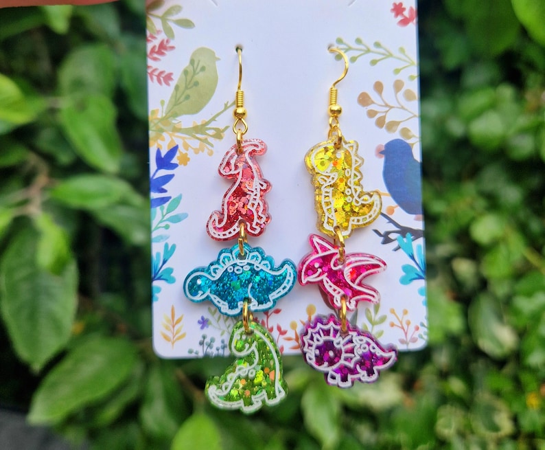 May include: Dangle earrings featuring colorful dinosaur shapes in red, yellow, blue, pink, and green with gold-colored hooks. The earrings have a glittery finish and are displayed on a white card with a floral design.